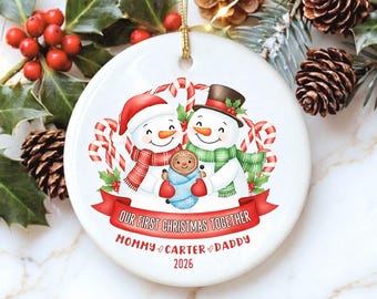 Op de afbeelding: Een rond wit keramieken ornament met twee sneeuwpoppen met sjaals en hoeden, die een ingebakerde peperkoekbaby vasthouden. Een rode banner bevat de tekst OUR FIRST CHRISTMAS TOGETHER, met de namen MOMMY, CARTER, DADDY en het jaar 2026.