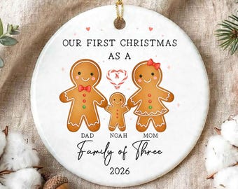 May include: A round white ceramic Christmas ornament featuring three gingerbread figures, labeled Dad, Noah, and Mom. The text reads Our First Christmas As A Family Of Three 2026. A candy cane heart sits between the figures, with small red hearts.