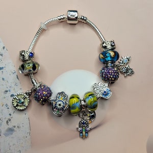 May include: A silver charm bracelet with a variety of colorful glass and metal charms, including a blue eye, a hamsa hand, and a purple flower.