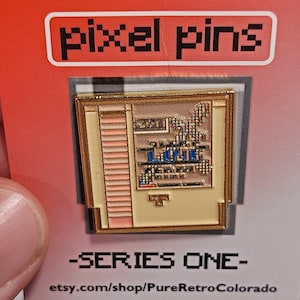 Puede incluir: Un pin de esmalte dorado con una imagen pixelada de una consola de videojuegos retro. El pin está en una tarjeta roja con el texto "pixel pins" y "SERIES ONE" en negro. La tarjeta también incluye la dirección del sitio web "etsy.com/shop/PureRetroColorado".