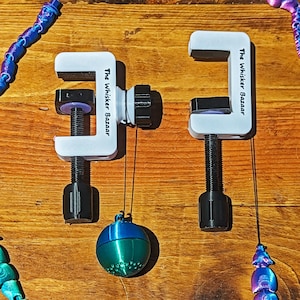 Puede incluir: Dos abrazaderas blancas y negras con el texto "The Whisker Bazaar". Cada abrazadera tiene un tornillo negro y un adorno colgante. Uno es una esfera azul y verde, el otro un diseño de cuentas azules y moradas.