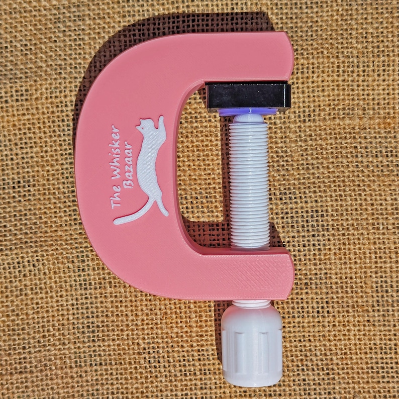 May include: A pink C-clamp with a white screw mechanism and a white handle. The clamp features the text "The Whisker Bazaar" and a white cat silhouette. The clamp is designed for crafts and DIY projects.