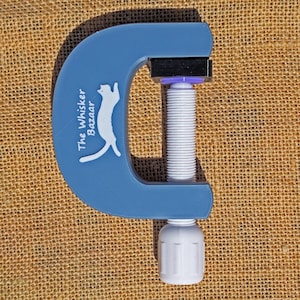 K&ouml;nnte beinhalten: Eine blaue C-Klemme mit wei&szlig;er Gewindestange und Griff. Die Klemme tr&auml;gt den Text "The Whisker Bazaar" und eine wei&szlig;e Katzen-Silhouette auf dem blauen Rahmen. F&uuml;r Bastelarbeiten und Heimwerkerprojekte.