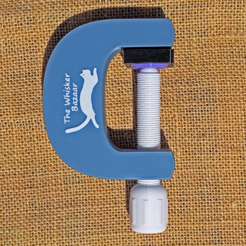 K&ouml;nnte beinhalten: Eine blaue C-Klemme mit wei&szlig;er Gewindestange und Griff. Die Klemme tr&auml;gt den Text "The Whisker Bazaar" und eine wei&szlig;e Katzen-Silhouette auf dem blauen Rahmen. F&uuml;r Bastelarbeiten und Heimwerkerprojekte.