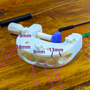 K&ouml;nnte beinhalten: Eine wei&szlig;e, 3D-gedruckte Klemme mit einer Schraube und einer blauen Kappe. Die Klemme hat mehrere L&ouml;cher und die Ma&szlig;e 8 mm, 18 mm und 13 mm sind sichtbar. Zwei schwarze St&auml;be befinden sich im Hintergrund.