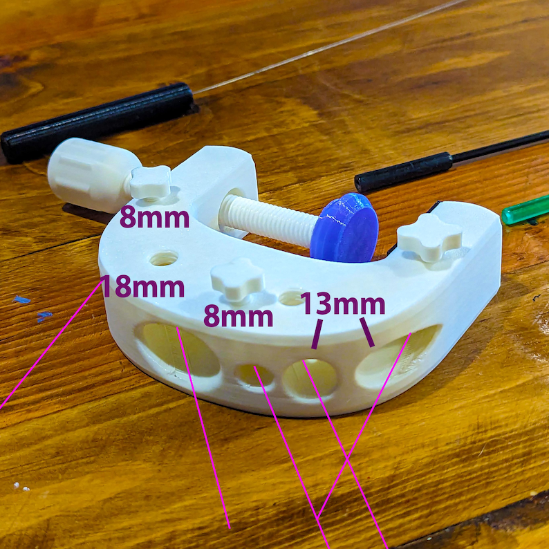 K&ouml;nnte beinhalten: Eine wei&szlig;e, 3D-gedruckte Klemme mit einer Schraube und einer blauen Kappe. Die Klemme hat mehrere L&ouml;cher und die Ma&szlig;e 8 mm, 18 mm und 13 mm sind sichtbar. Zwei schwarze St&auml;be befinden sich im Hintergrund.