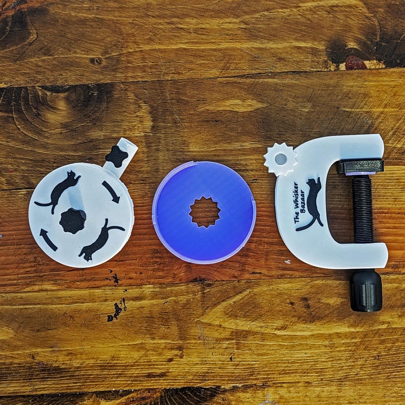 May include: Three white and blue cat-themed tools on a wooden surface. The tools include a circular object with cat silhouettes, a blue circular object with a star-shaped center, and a clamp with the text "The Whisker Bazaar" and a cat silhouette.