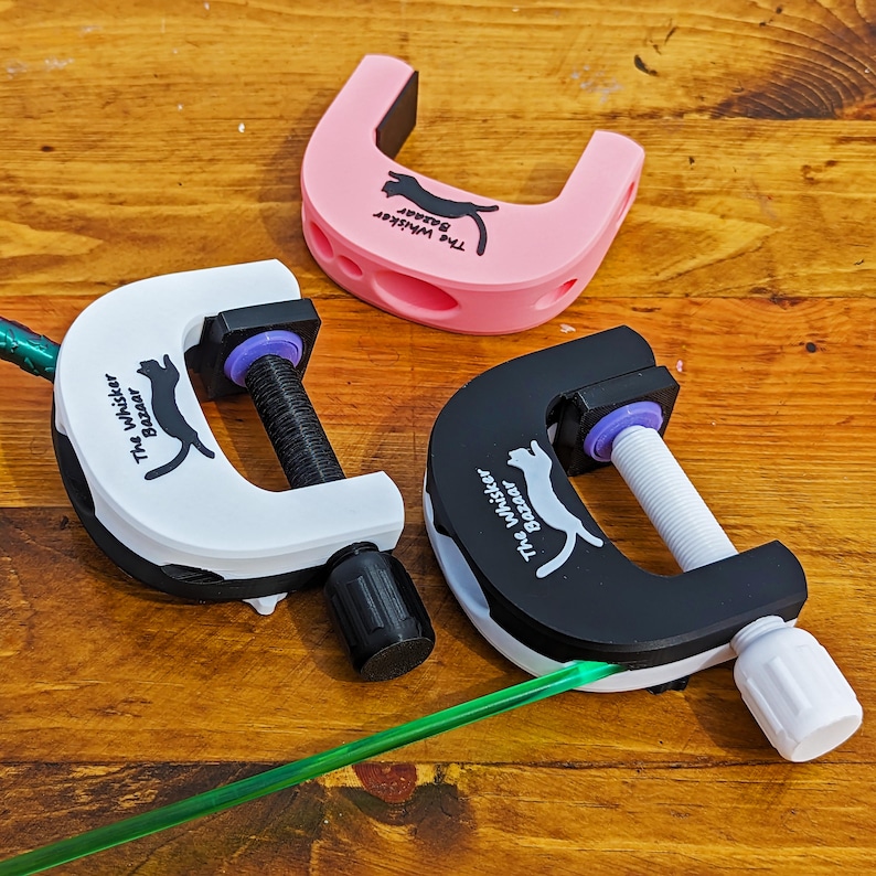 K&ouml;nnte beinhalten: Drei C-f&ouml;rmige Katzen-Spielzeuge in Rosa, Wei&szlig; und Schwarz. Jede Klemme hat einen Schraubmechanismus und den Text "The Whisker Bazaar" mit einer Katzen-Silhouette. Die Spielzeuge liegen auf einer Holzoberfl&auml;che.