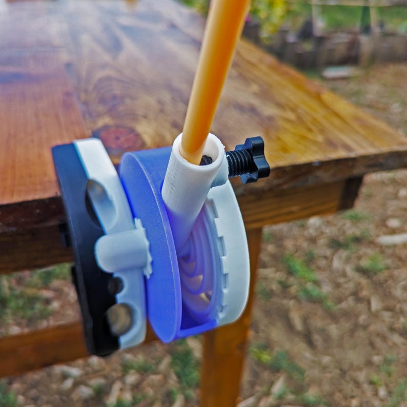 May include: A 3D-printed device with a blue, white, and black color scheme, featuring a circular design with a central tube and a black adjustment knob. An orange tube is inserted into the device. The device is clamped to a wooden surface.