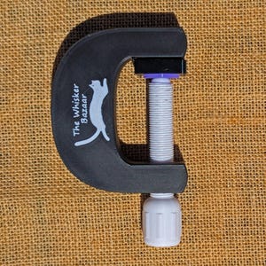 May include: Black C-clamp with a white threaded rod and a white handle. The clamp has the text "The Whisker Bazaar" and a cat logo printed on the side. The clamp is made of a matte black material.
