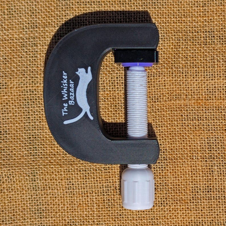May include: Black C-clamp with a white threaded rod and a white handle. The clamp has the text "The Whisker Bazaar" and a cat logo printed on the side. The clamp is made of a matte black material.