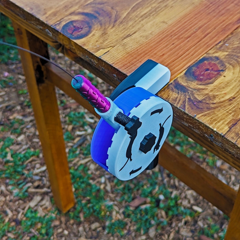 May include: A blue and white cable winder attached to a wooden table. The winder has a purple handle and a black knob. The table is made of wood and has a natural finish. The winder is designed to manage cables.