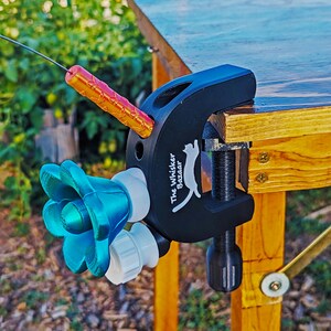 K&ouml;nnte beinhalten: Eine schwarze Klemme mit einem silbernen Schraubmechanismus ist an einer Holzoberfl&auml;che befestigt. Ein blaues bl&uuml;tenf&ouml;rmiges Objekt und ein zylindrisches Objekt in Orange und Rosa sind an der Klemme befestigt. Der Text "The Whisker Bazaar" ist auf die Klemme gedruckt.