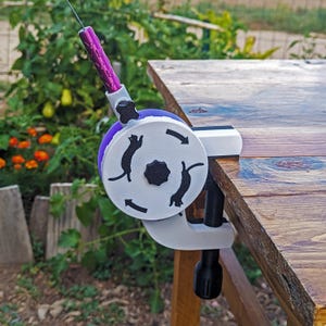 May include: A purple and white cat toy with a clamp, designed to attach to a table. The toy features a circular design with black cat silhouettes and directional arrows. The handle is purple with a textured grip.