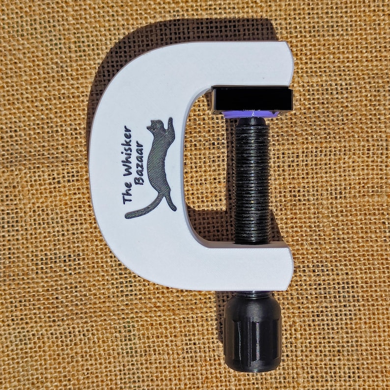 May include: A white C-clamp with a black threaded rod and handle. The clamp has the text "The Whisker Bazaar" and a cat silhouette. The clamp is on a tan burlap surface.