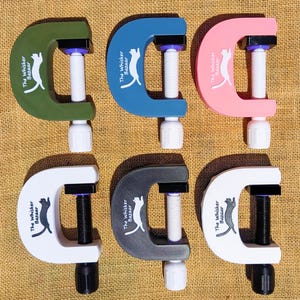 K&ouml;nnte beinhalten: Sechs C-Klemmen in verschiedenen Farben: Gr&uuml;n, Blau, Rosa, Wei&szlig;, Grau und Wei&szlig;. Jede Klemme hat eine wei&szlig;e Schraube und einen schwarzen Griff. Der Text "The Whisker Bazaar" und eine Katzen-Silhouette sind aufgedruckt.