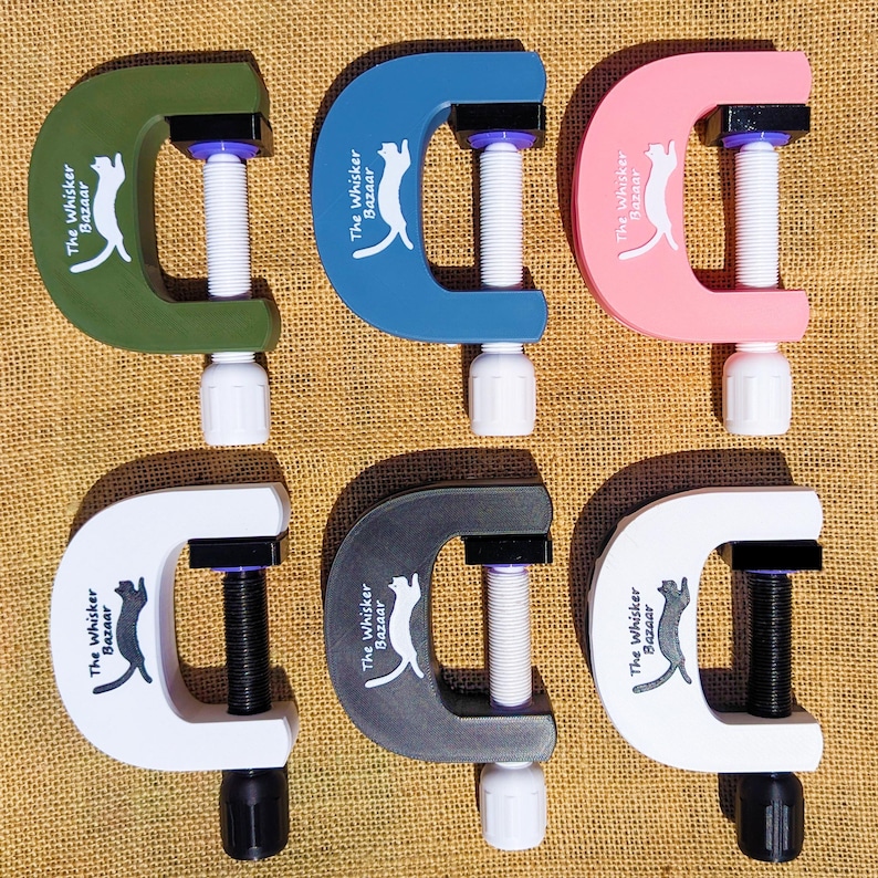K&ouml;nnte beinhalten: Sechs C-Klemmen in verschiedenen Farben: Gr&uuml;n, Blau, Rosa, Wei&szlig;, Grau und Wei&szlig;. Jede Klemme hat eine wei&szlig;e Schraube und einen schwarzen Griff. Der Text "The Whisker Bazaar" und eine Katzen-Silhouette sind aufgedruckt.