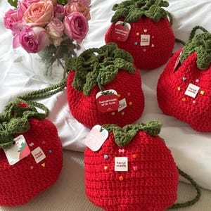 Puede incluir: Una colección de bolsos hechos a mano con forma de fresa de ganchillo rojo con la parte superior de hojas verdes. Cada bolso tiene un cierre de cordón y una correa. Pequeñas etiquetas dicen "handmade with love" y "hand made". Un ramo de rosas rosas está en el fondo.