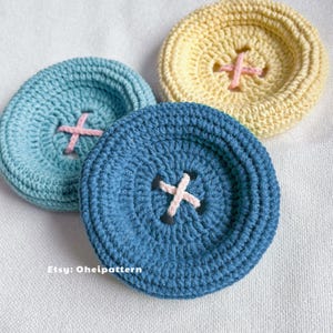 May include: Three handmade crochet coasters in shades of blue and yellow, resembling large buttons. Each coaster features a cross-stitch design in a contrasting color. The Etsy shop name "Oheipatter" is visible in the bottom left corner.