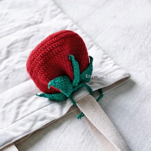 Pode incluir: Um morango de crochê feito à mão, com um corpo vermelho vibrante e folhas verdes. A fruta artesanal está colocada em uma sacola branca. A imagem é bem iluminada, mostrando a textura da linha e os detalhes do trabalho de crochê.