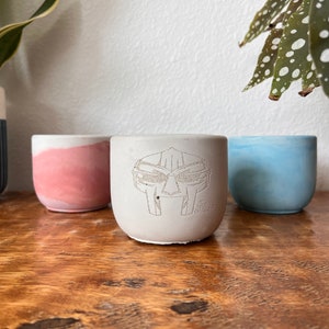 Könnte beinhalten: Drei kleine Beton-Pflanztöpfe, einer mit rosa Glasur, einer mit blauer Glasur und einer mit weißer Glasur und einer schwarz-weißen Grafik eines Helms mit Gesichtsmaske. Die Töpfe stehen auf einer Holzoberfläche.