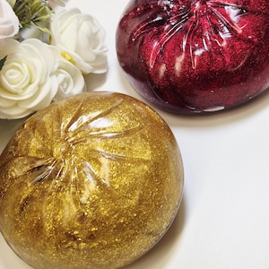 Könnte beinhalten: Zwei runde, glatte, kuppelförmige Objekte mit glänzender Oberfläche. Ein Objekt ist dunkelrot mit eingearbeitetem Glitzer, das andere ist goldfarben mit eingearbeitetem Glitzer. Beide weisen einen Mittelpunkt mit strahlenförmigen Linien auf.