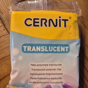 May include: A yellow package of translucent polymer clay by Cernit. The package has the brand name Cernit and the word Translucent printed on it. The package also includes the text "Pâte polymère translucide", "Translucent polymer clay", "Transluzente Polymermasse", "Pasta Polimerica traslucida", and "Arcilla polimérica translúcida".