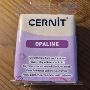 Może przedstawiać: Opakowanie białej gliny polimerowej z ceramicznym wykończeniem. Opakowanie jest oznaczone "CERNIT OPALINE" i zawiera tekst "Pâte polymère finition céramique, Polymer clay with ceramic finish, Polymermasse mit keramik-ähnliches Finish, Pasta Polimerica con effetto ceramica, Arcilla polimérica con acabado de cerámico."