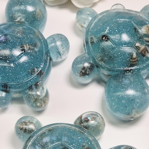 Könnte beinhalten: Mehrere blaue Schildkrötenfiguren aus Kunstharz mit eingebetteten Muscheln und Glitzer. Die Schildkrötenpanzer haben ein geometrisches Muster. Die Figuren befinden sich auf einer weißen Oberfläche, mit weißen Blumen im Hintergrund.
