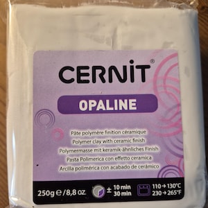 Może przedstawiać: Biały blok gliny polimerowej CERNIT OPALINE z fioletową i różową etykietą. Etykieta zawiera tekst "CERNIT OPALINE" i informacje o glinie, w tym jej zastosowanie, czas pieczenia i temperaturę. Opakowanie jest oznaczone jako 250g/8,8 oz.