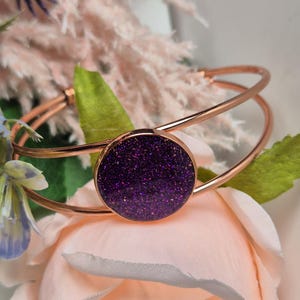 Op de afbeelding: Een roségouden armband met een ronde paarse glitterbedel. De armband is gemaakt van twee dunne metalen banden.