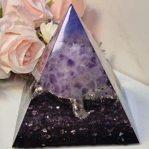 Könnte beinhalten: Eine klare Glaspyramide gefüllt mit violetten Amethystkristallen und Glitzer. Die Pyramide steht auf einer weißen Oberfläche.