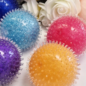 Può includere: Una collezione di palline antistress colorate e appuntite nei toni del blu, turchese, rosa, arancione e viola. Le palline hanno una superficie strutturata e sono disposte con rose bianche e rosa sullo sfondo.