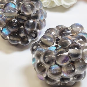 Könnte beinhalten: Zwei kugelförmige Perlen mit traubenartiger Textur aus durchscheinendem dunkelgrauem Harz. Jede Perle enthält eingebettete irisierende blaue und violette Glitzerpartikel, die das Licht einfangen und einen schimmernden Effekt erzeugen.