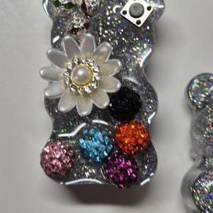 Pode incluir: Um amuleto de urso de resina com glitter prateado com uma flor branca, uma pérola e pedras brilhantes coloridas. O amuleto de urso tem um fundo de glitter prateado e um pequeno ímã quadrado na parte de trás.