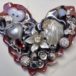 Pode incluir: Uma peça de resina em forma de coração com um fundo marmorizado vermelho e branco. É decorada com vários enfeites, incluindo botões brancos e prateados, contas, strass e uma flor branca com um centro prateado.