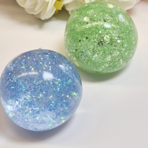 Könnte beinhalten: Zwei kugelförmige Objekte mit einer glatten, glänzenden Oberfläche. Eine Kugel ist hellblau und mit feinen, irisierenden Glitzerpartikeln gefüllt. Die zweite Kugel ist hellgrün und enthält eine Mischung aus feinen und groben silberfarbenen Glitzerflocken.