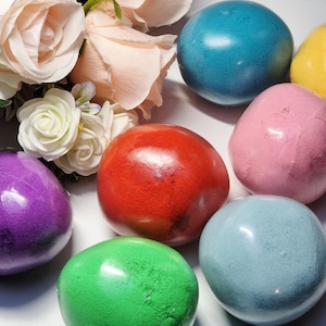 Könnte beinhalten: Eine Auswahl an farbenfrohen, glatten, kugelförmigen Objekten in den Farbtönen Rot, Grün, Blau, Lila, Rosa und Gelb. Die Objekte sind um einen Strauß weißer und pfirsichfarbener Rosen angeordnet.