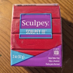 Può includere: Una confezione rossa di argilla da forno Sculpey III. La confezione è etichettata con il nome del marchio Sculpey e il nome del prodotto Sculpey III. La confezione include anche il peso dell'argilla, 2 once (57 grammi), e le istruzioni per cuocere l'argilla.