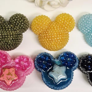 Könnte beinhalten: Eine Sammlung bunter, Mickey Mouse-förmiger Harzdekorationen. Die Dekorationen sind in verschiedenen Farben erhältlich, darunter Gold, Rosa, Blau und Schwarz, mit sternförmigen Akzenten. Die Dekorationen befinden sich auf einer weißen Oberfläche.