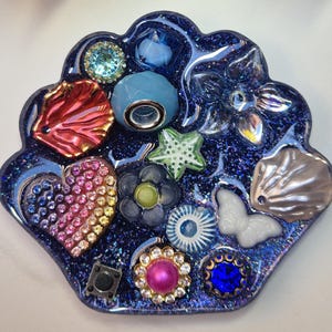Pode incluir: Uma peça decorativa de resina azul brilhante com vários enfeites embutidos. Apresenta uma concha vermelha, um coração arco-íris, uma borboleta branca e várias contas e flores com tons de joia. A peça tem uma borda ondulada.