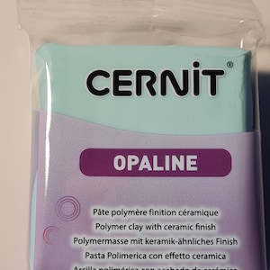 Może przedstawiać: Opakowanie niebieskiej gliny polimerowej z ceramicznym wykończeniem. Opakowanie jest oznaczone "CERNIT® OPALINE" i zawiera tekst "Pâte polymère finition céramique, Polymer clay with ceramic finish, Polymermasse mit keramik-ähnliches Finish, Pasta Polimerica con effetto ceramica, Arcilla polimérica con acabado de cerámico."