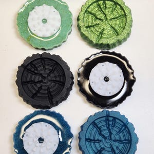 Peut inclure: Six dessous de verre circulaires en résine disposés en deux colonnes. Trois dessous de verre présentent un centre blanc texturé en forme d'engrenage sur des bases à paillettes vertes, noires et bleues. Les trois autres arborent un motif de cernes.