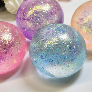 Könnte beinhalten: Eine Ansammlung von vier durchscheinenden, kugelförmigen Objekten mit einer glatten, glänzenden Oberfläche. Jede Kugel ist mit irisierenden Glitzerpartikeln in Rosa, Lila, Blau und Pfirsich gefüllt, was einen reflektierenden Glanzeffekt erzeugt.