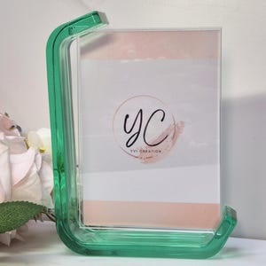 Può includere: Una cornice per foto in acrilico trasparente con un supporto curvo verde brillante. La cornice contiene un'immagine con uno sfondo rosa chiaro e bianco, con il testo "yc" all'interno di un cerchio, con "YVI CREATION" sotto.