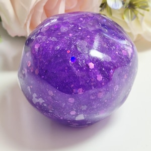 Könnte beinhalten: Ein abgerundetes, durchscheinendes violettes Objekt mit einer glatten, glänzenden Oberfläche. Das Material ist mit verschieden großen, irisierenden und holografischen Glitzerpartikeln durchsetzt, die das Licht reflektieren und einen funkelnden Effekt erzeugen.
