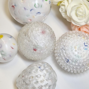 Peut inclure: Un ensemble de quatre objets sphériques à base blanche avec des taches multicolores semblables à des confettis. Deux sphères ont des surfaces lisses, tandis que les deux autres présentent des extérieurs texturés et bosselés. Des roses artificielles crème sont visibles.