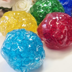 Op de afbeelding: Een verzameling van vijf doorschijnende, bolvormige stressballen met een getextureerd, hobbelig oppervlak. De ballen zijn gekleurd in heldergeel, levendig rood, lichtblauw, donkergroen en diepblauw, elk met een glanzende, reflecterende afwerking.