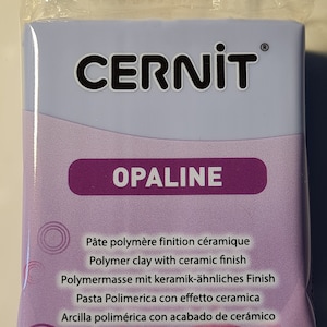 Może przedstawiać: Opakowanie białej gliny polimerowej z fioletową etykietą, na której widnieje napis "CERNIT OPALINE" oraz lista właściwości gliny w kilku językach. Glina jest przeznaczona do rękodzieła i ma ceramiczne wykończenie.