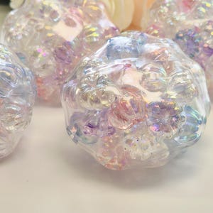 Könnte beinhalten: Nahaufnahme von mehreren irisierenden, klaren, blütenförmigen Perlen. Die Perlen haben eine strukturierte Oberfläche und reflektieren einen Regenbogen aus Farben, darunter Rosa, Lila und Blau. Die Perlen sind zusammengefasst und erzeugen einen funkelnden Effekt.